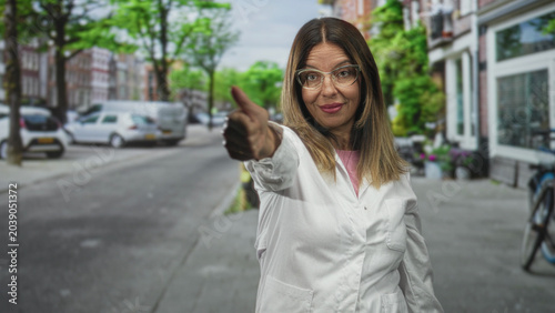 Canvas-taulu Woman doctor in labcoat gives thumbs up showing thumb and smiling while standing on a street sidewalk near storefronts, cars and bicycle; confidence care