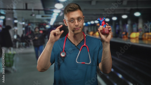 Obraz na płótnie Man cardiologist in scrubs holds anatomical heart model aloft with right hand while stethoscope hangs around his neck in airport; confidence cardiac education care