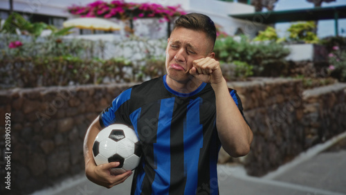 Canvas Print Man holding soccer ball, pouting and rubbing eye with knuckle on paved street beside stone wall and flowering planters; playful disappointment