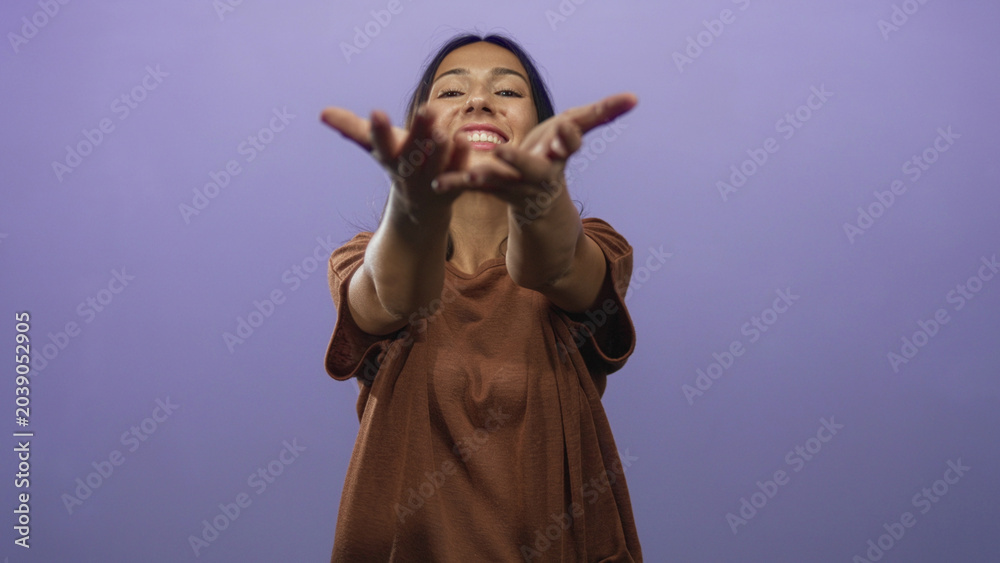 Obraz premium Young hispanic brunette woman with outstretched hands reaching toward camera in a purple studio; warm welcoming joy.