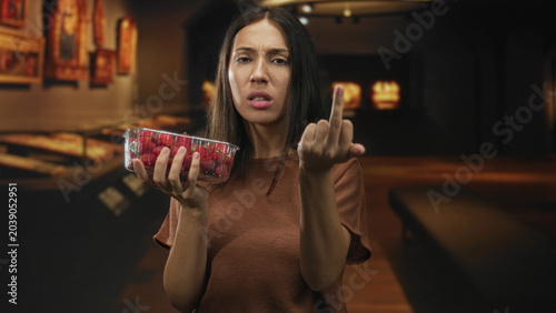 Valokuva Hispanic woman holds clear container of strawberries and shows middle finger in art gallery; defiance attitude rebellion