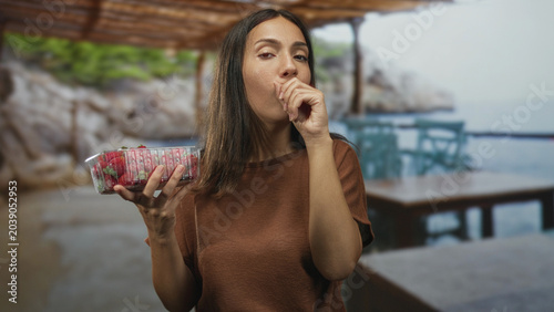 Photos Young brunette woman holding plastic punnet of strawberries and covering mouth while coughing at beach cafe terrace by the sea; discomfort