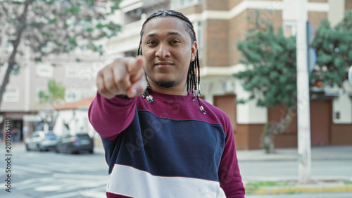 Slika na platnu Man giving thumb down gesture on street in front of apartment building while wearing burgundy and navy sweater and braided hair; disapproval casual