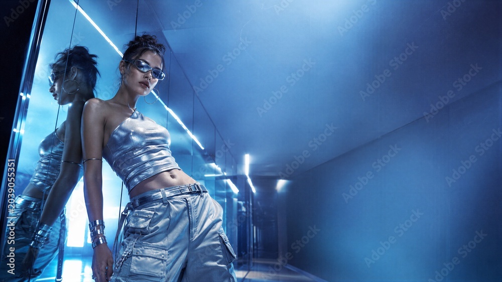 Naklejka premium Woman leaning against mirror wearing stylish metallic y2k clothing. Futuristic fashion model with sunglasses in blue neon light studio. Retro aesthetic style from the early two thousands