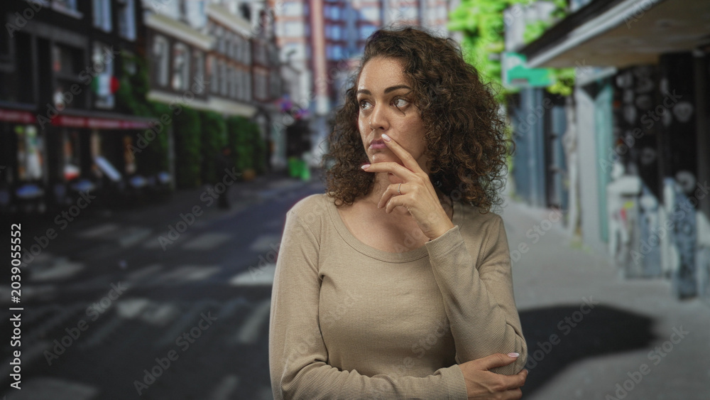Fototapeta premium Woman with hand on chin at a city street crosswalk, neutral expression and folded arm across body, visible ring on finger; quiet contemplation.