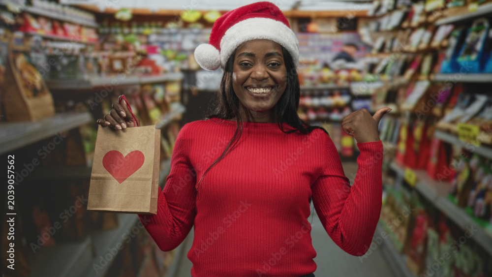 Obraz premium Woman in santa hat and red sweater holding a paper gift bag with heart while pointing thumb toward aisle in a building; holiday joy.