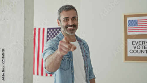 Fotografie Man points finger at camera beside 'every vote counts' poster and us flag in a voting building; civic duty determination