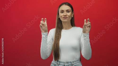 Foto Woman making air quotes with both hands, showing fingers and palms, neutral skeptical expression, long hair, wearing white top and jeans, posed against a red studio wall; skepticism