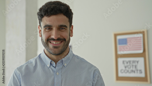 Fotografie Man smiling with visible face and beard, looking at camera in a building with an american flag poster and 'every vote counts' sign; civic duty hopeful