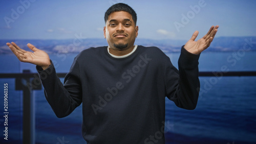 Quadro su tela Man palms up shrugging on a building balcony by the sea with casual expression and blue ocean vista behind him; subtle uncertainty
