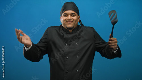 Foto Man chef in black uniform and skullcap holding a spatula, shrugging shoulders and smiling in studio midshot; cheerful hospitality