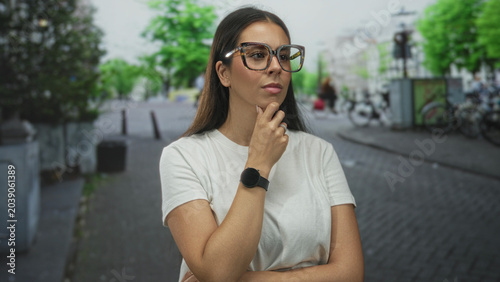Konstfotografi Woman with long brunette hair wearing white t shirt and oversized glasses, smartwatch visible, hand on chin on a cobblestone street with bicycles nearby; contemplation