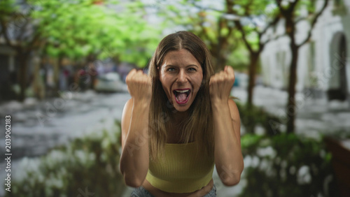 Фотография Young woman with bare midriff and bellybutton visible, fists raised above head in yellow crop top on a busy tree lined street, laughing and cheering with triumphant grin; joy victory