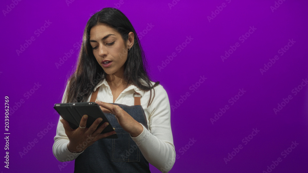 Naklejka premium Woman in a denim apron and white shirt tapping a tablet with fingers while reviewing an order list in a purple studio; concentration productivity.
