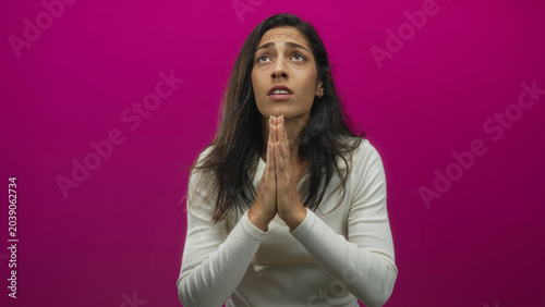 Canvas Print Young hispanic woman praying with clasped hands in studio against magenta backdrop; hopeful plea for comfort