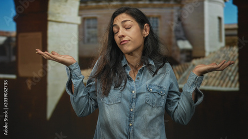 Konstfotografi Young hispanic woman with hands raised palms out shrugging on old town street, wearing denim shirt and rings, casual shrug and slight smirk; indifference