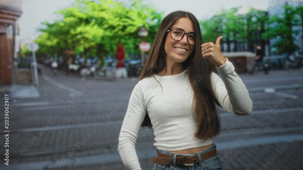 Fototapeta premium Young hispanic woman wearing glasses smiles and gives thumbs up with bare hands on an urban street with trees and bikes; confidence positivity.
