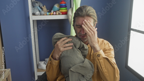 Fotografia Man cradling wrapped baby against his chest, hands visible and hand to face gesture beside a crib in a nursery; fatigue tenderness