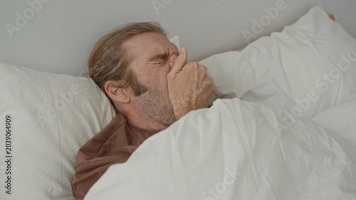 Photos Man in bed covering mouth with hand while yawning, pillow and white duvet visible, wooden headboard in building; morning fatigue