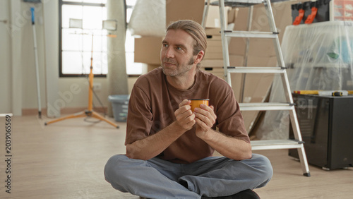 Photos Man holding mug with both hands seated crosslegged in building beside ladder, stacked boxes and covered furniture; contemplation renovation pause