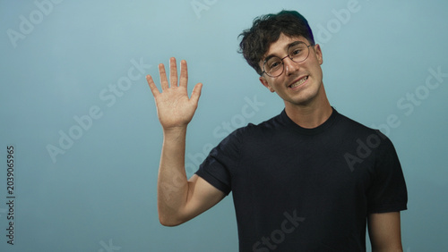 Photos Young hispanic man raising right hand with open palm to camera in light blue studio, wearing black t shirt and round glasses; hesitant greeting