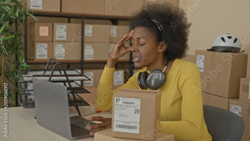 Obraz na plátně Woman rubbing temple with hand near parcel and laptop amid stacked delivery boxes in a packing building with apparent headache; small business fatigue