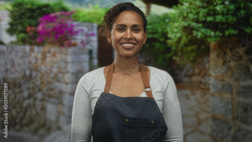 Konstfotografi Woman in denim apron smiling with visible teeth while standing on a stone street beside flowering wall and greenery; contentment hospitality