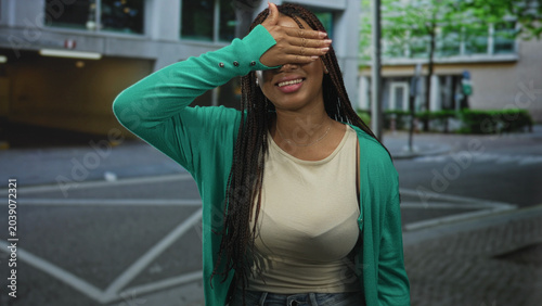 Bild auf Leinwand Woman in green cardigan covering eyes with hand and smiling on urban street near building crosswalk; shy joy