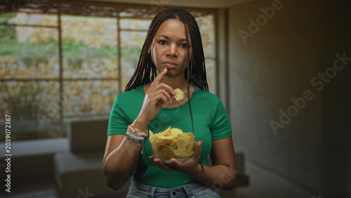 фотографія Woman holding glass bowl of chips with finger to lips for silence in a building living room; thoughtful pause