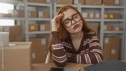 Принти на полотні Young woman with glasses resting head on hand at packing table with laptop and cardboard boxes on shelves in a building; tired monotony