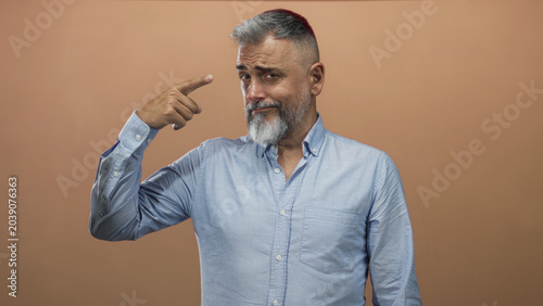 Photography Man pointing finger to temple in studio with peach backdrop and light blue shirt, gray beard and raised eyebrow; wry humor skepticism