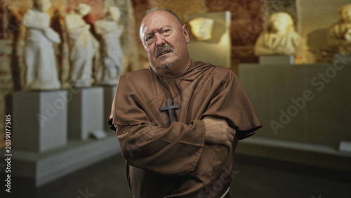 Cuadro en lienzo Man monk in brown robe and wooden cross clutches his arm, grimaces while standing among marble statues inside a museum building; suffering reflection