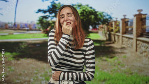 Billede på lærred Woman biting fingernail with hand to mouth, eyes closed, striped top visible, standing near stone fence and park lawn on street; nervousness vulnerability