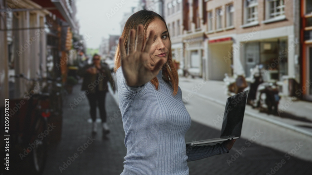 Fototapeta premium Woman holds laptop in one hand and extends palm forward in a crowded street, gaze stern and dismissive; defiance privacy.