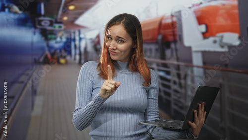 Photos Young redhead woman points finger while holding laptop in ship corridor with orange lifeboat and railings visible, looking skeptical; warning doubt