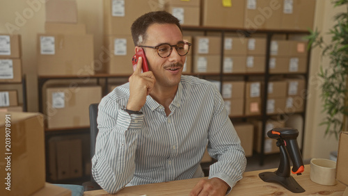 Man with mustache holding smartphone to ear and smiling at desk with boxes and barcode scanner in building; entrepreneurial calm.