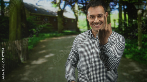 Fotografija Man with moustache and checkered shirt showing middle finger and smiling while standing on a forest path near a cottage; defiance