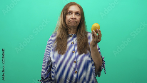Quadro su tela Woman in striped blouse holds a lemon aloft in her right hand in a green studio, looking unimpressed; resignation reflection