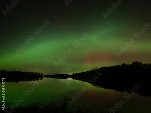 Northern lights of red and green fill the sky and reflect in the waters of a lake below
