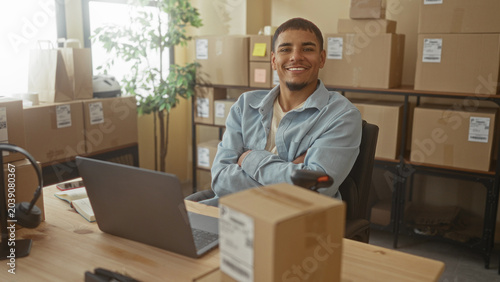 Bild auf Leinwand Man types on laptop amid stacked shipping boxes, holds barcode scanner and crosses arms in some frames in small business packaging studio; entrepreneurial confidence