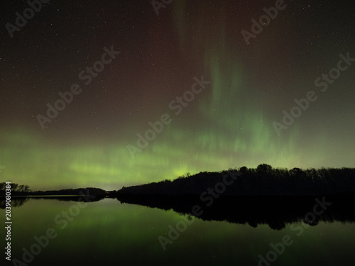 Northern lights of red and green fill the sky and reflect in the waters of a lake below