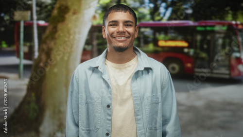 Photos Man smiling with visible face in a light blue denim shirt standing beside a large tree on a street near a red bus and sidewalk; warm optimism