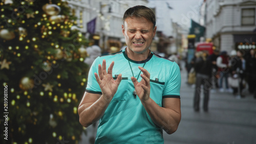 Foto Man referee with whistle wincing face and hands raised near chest on street christmas market; discomfort caution