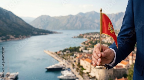 Businessman holding a miniature flag of Montenegro overlooking the blurry Kotor bay, mountains, and coastal town