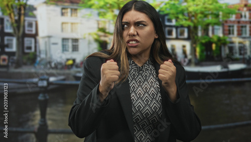фотографія Woman grimaces and clenches fists on a canal street by residential buildings and a railing, face contorted and nose piercing visible; frustration resolve