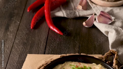 A rustic close up shot of a creamy homemade hummus bowl garnished with olive oil, chili slices, and fresh herbs, set on a dark wooden table with raw garlic and chili peppers in the background.