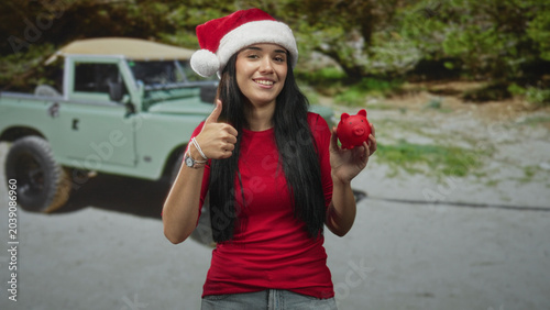 Fotografia Woman holding red piggy bank and giving thumbs up while wearing santa hat in forest beside a parked jeep; holiday savings joy