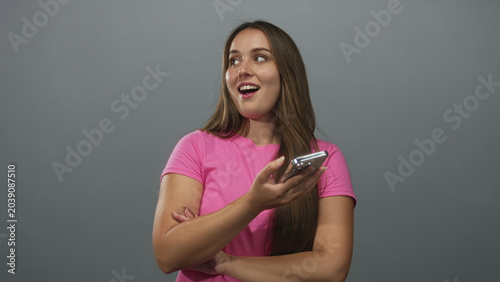 Foto Woman in pink t shirt holding smartphone, smirking and glancing sideways in grey studio; amused curiosity