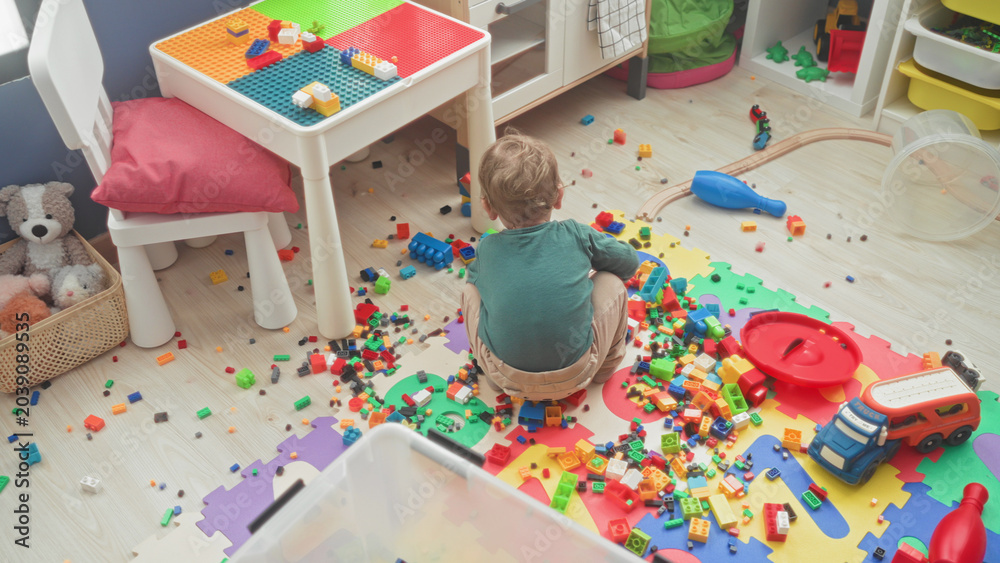 Fototapeta premium Boy sitting on foam puzzle mat reaching for scattered toy blocks and red toy truck in a cluttered play area inside a building; playful curiosity.