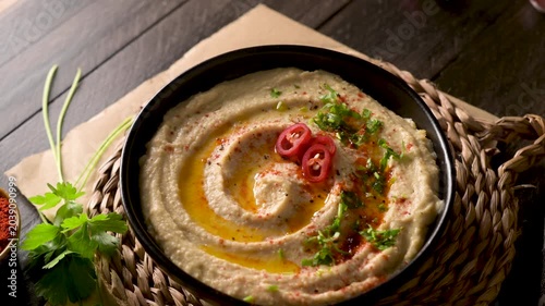 A close-up shot of creamy hummus in a dark bowl, drizzled with olive oil and topped with fresh chili slices and herbs. The rustic composition features soft, warm lighting on a dark wooden background.
